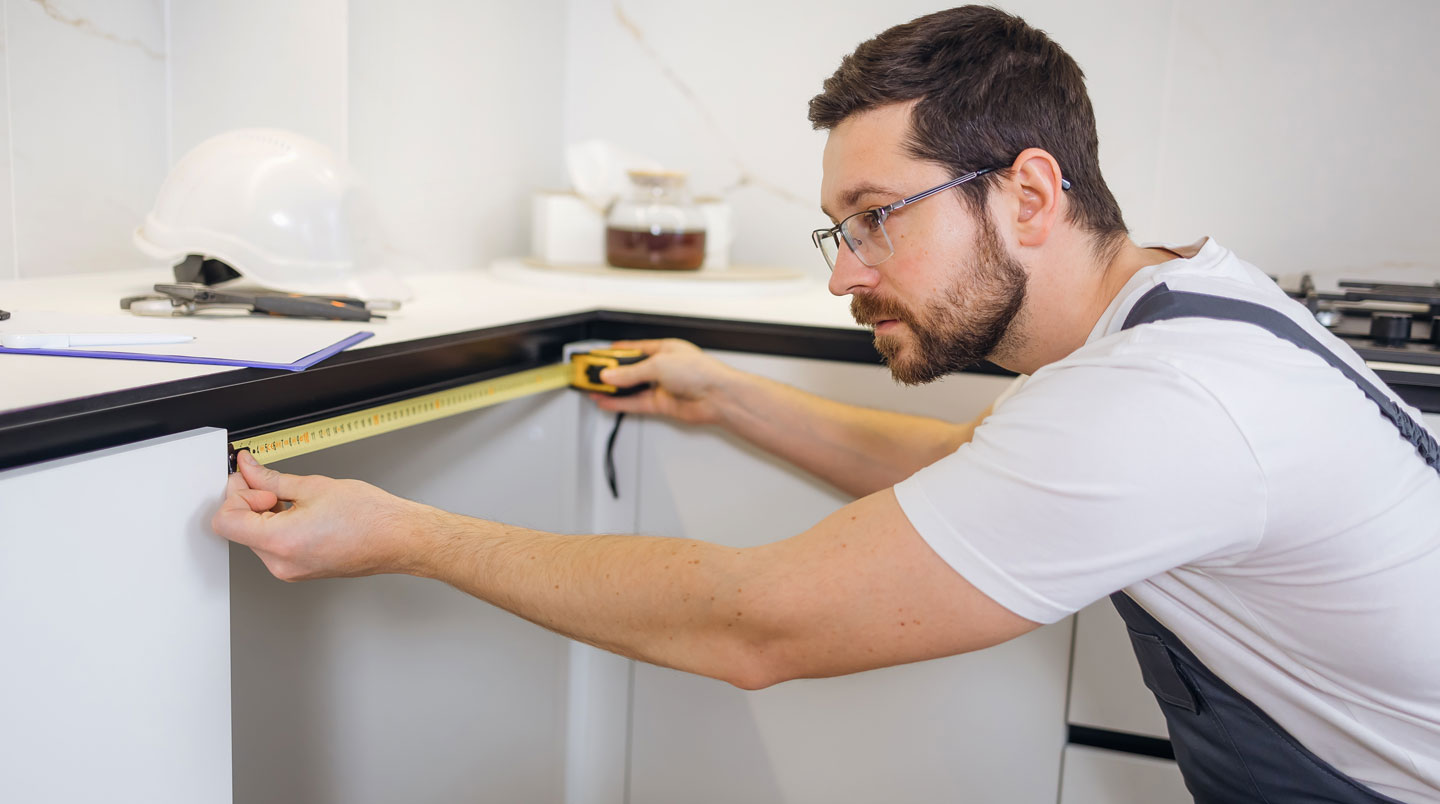 Main tenant un mètre ruban métallique mesurant la largeur d'une niche d'encastrement vide sous plan de travail dans un mobilier de cuisine moderne